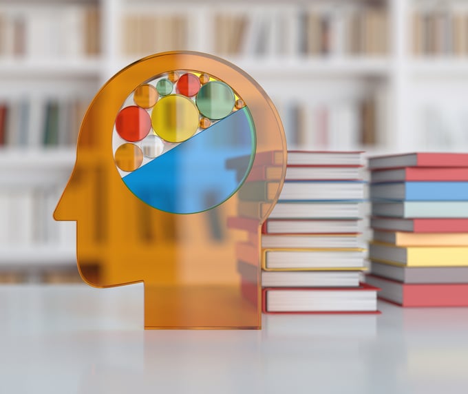Transparent orange silhouette of a human head with a colorful abstract brain made of circles and geometric shapes, set on a table beside stacks of books with blurred bookshelves in the background.