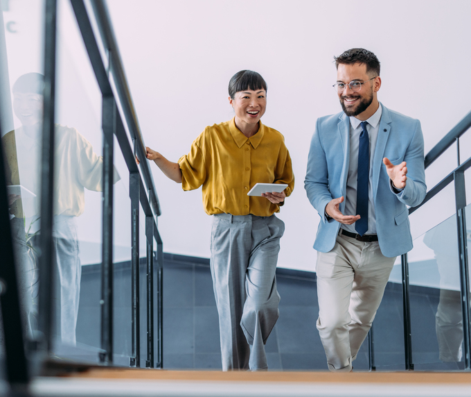 Two business professionals smiling and talking while walking up a modern office staircase, with one holding a tablet and the other gesturing as they converse.