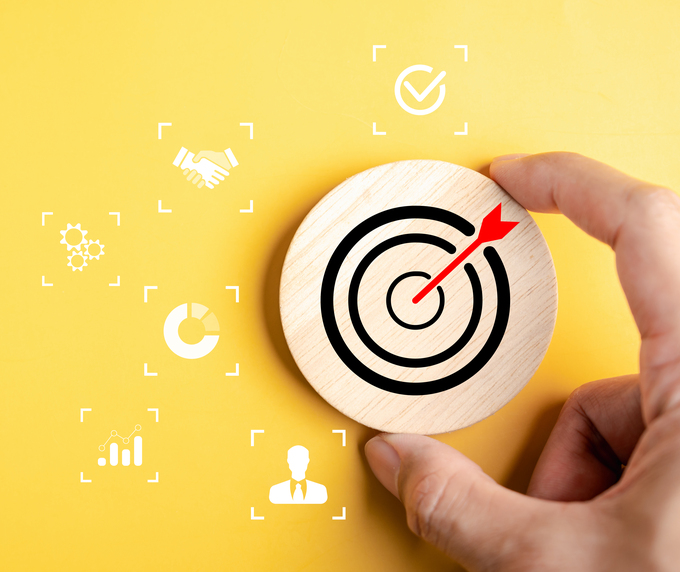 Hand placing a round wooden token with a bullseye and red arrow on a yellow background, surrounded by white business icons representing goals, partnership, analytics, settings, performance, and leadership.